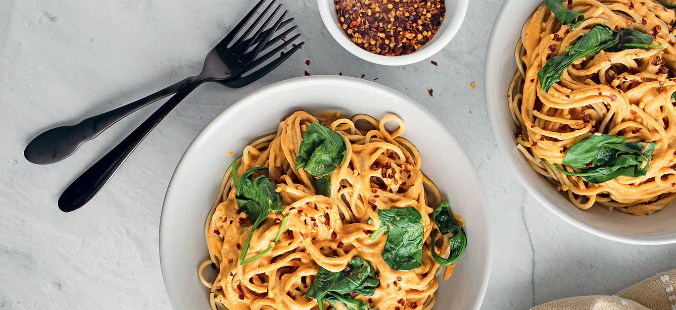 Two bowls of Brandi Doming's Harissa Almond Pasta with Spinach on a marble table with two forks and a small bowl of red pepper flakes