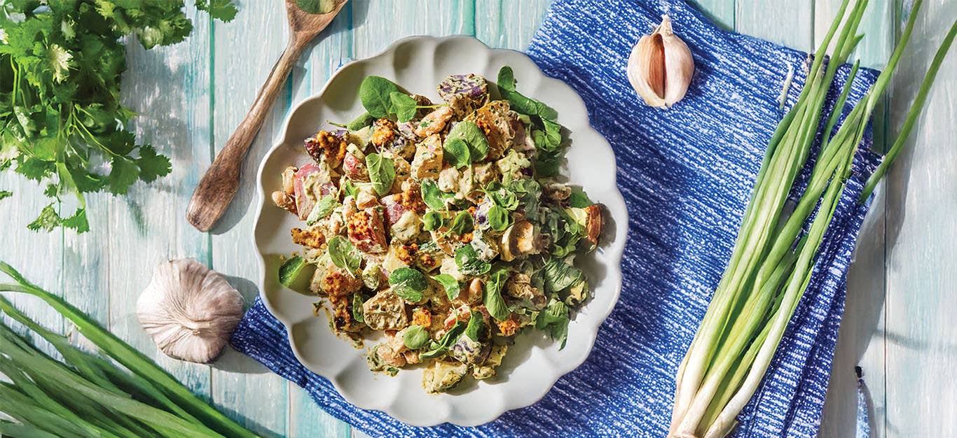 New Potato Salad with Herbed Vinaigrette on a plate on a blue-washed wooden table surrounded with a bulb of garlic, a bunch of spring onions, and a bunch of flat leaf parsley