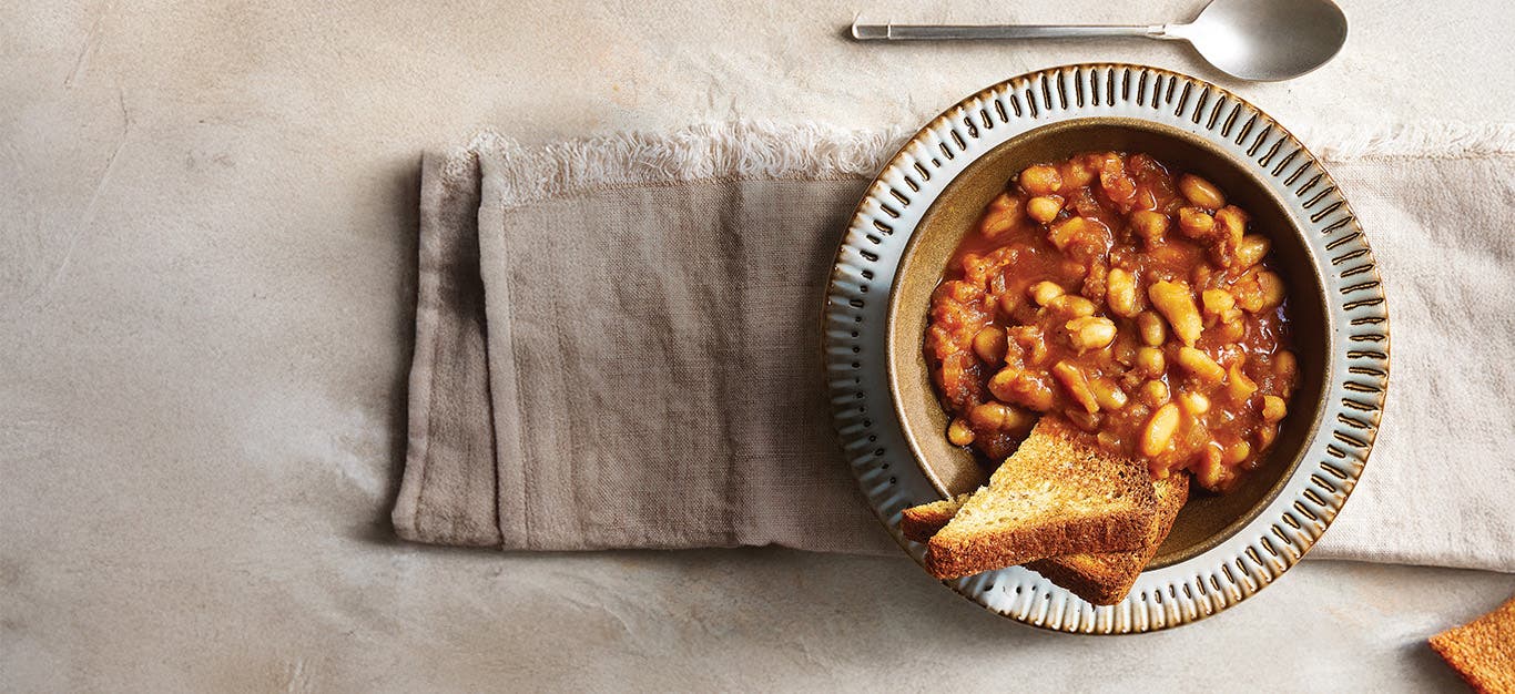 A bowl of baked beans made in a slow cooker, in a serving bowl with a slice of whole grain bread on the side