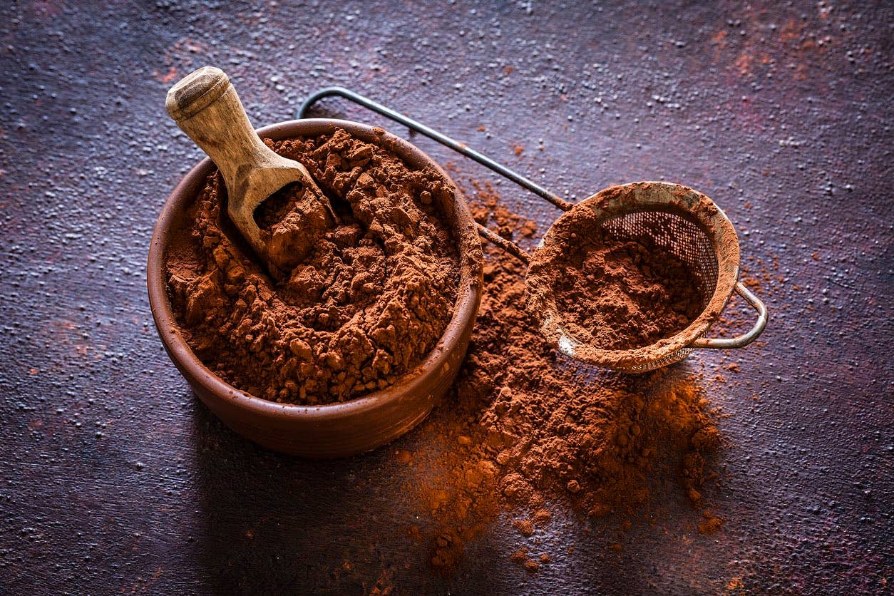 Brown bowl filled with cocoa powder shot on rustic table. Beside the bowl is a sieve also with cocoa powder.