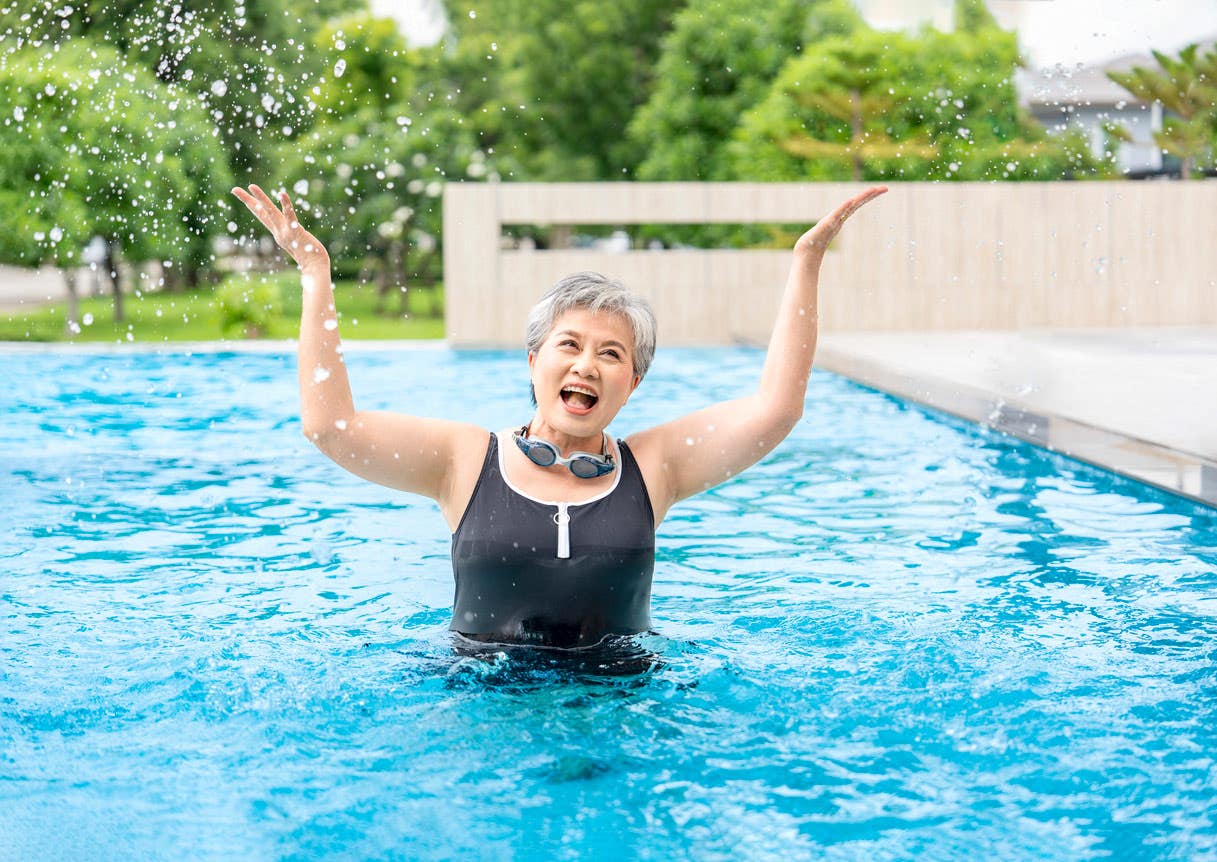 A senior woman stands in a pool outside looking joyous with her arms raised upward, wearing an athletic swimsuit and goggles around her neck